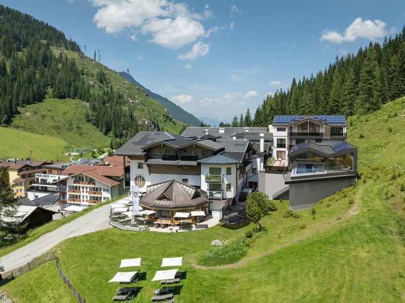 Hotelgebäude in einem grünen Tal mit Bergen im Hintergrund und Sonnenliegen auf der Wiese.