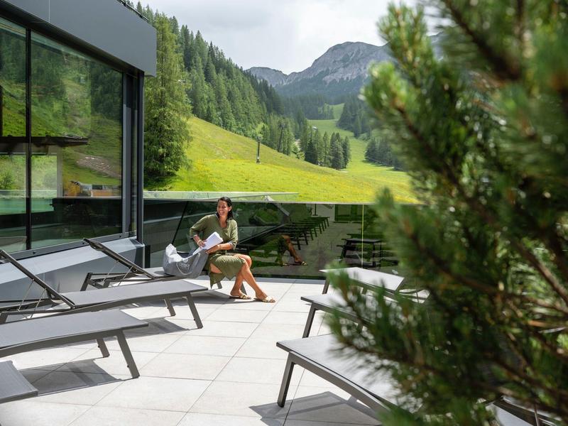 Terrasse mit Liegestühlen und Frau vor moderner Glasfassade mit Berglandschaft im Hintergrund