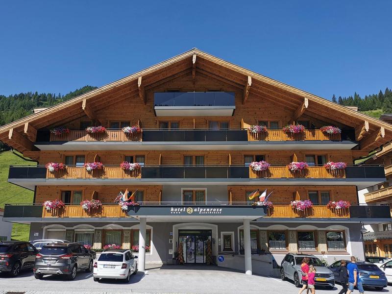 Mehrstöckiges Chalet-Hotel mit Blumen an Balkonen und geparkten Autos davor bei klarem Himmel.