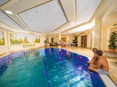 Innenpool mit Kindern, die am Beckenrand sitzen und springen, umgeben von hellen Wänden und Pflanzen.