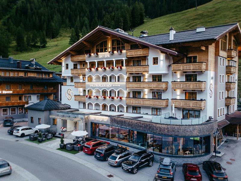 Mehrstöckiges Hotel in alpiner Landschaft mit zahlreichen geparkten Fahrzeugen davor.