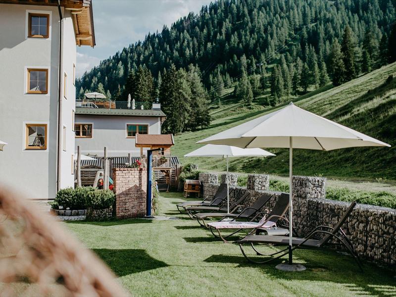 Grüner Garten mit Sonnenschirm und Liegestühlen neben einem Alpenhotel vor bewaldetem Berg.