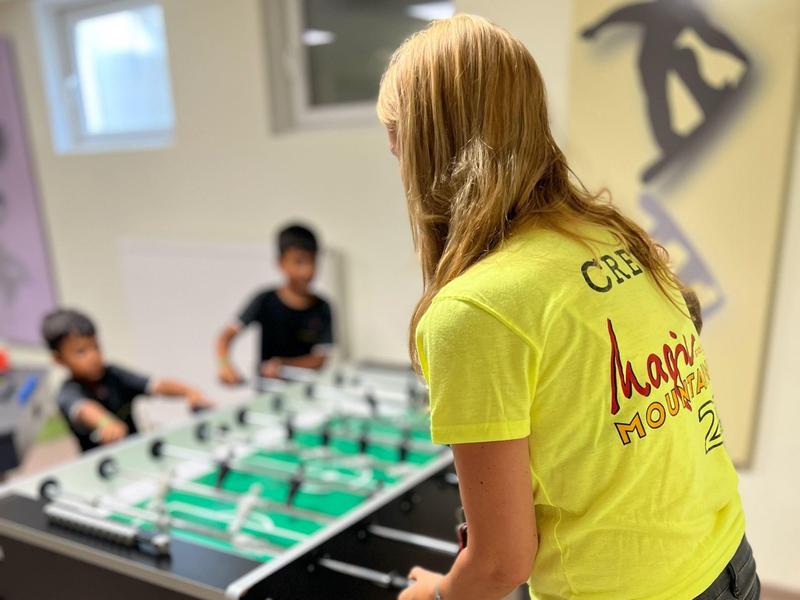 Person in gelbem T-Shirt spielt Tischfußball mit drei anderen in einem Raum