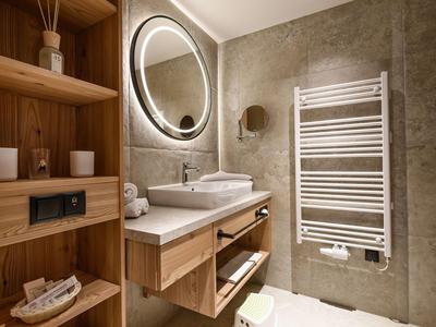 Modernes Badezimmer mit Holzregalen, Waschbecken, rundem beleuchtetem Spiegel und Handtuchheizung.