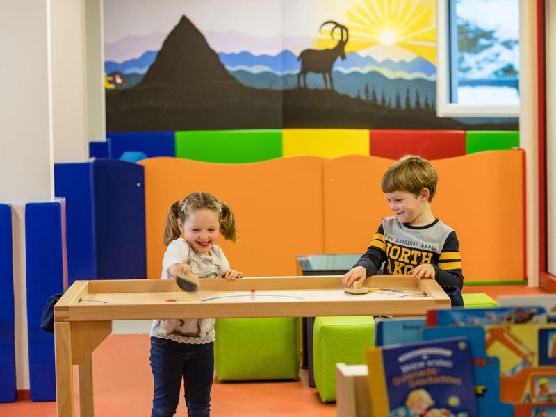 Zwei Kinder spielen an einem Holztisch in einem farbenfrohen Raum mit Sitzkissen und Bergwandbild.