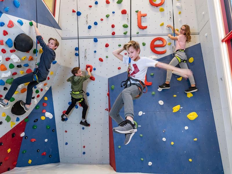 Vier Kinder klettern an einer bunten Indoor-Kletterwand in einer hellen Halle.