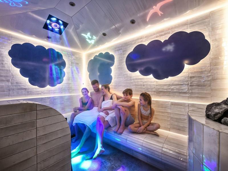 Fünf Personen sitzen in einer beleuchteten Wellness-Sauna mit Wolkenmotiven an der Wand.