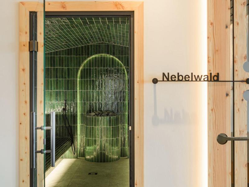 Blick durch eine Holztür auf einen grünen, mit Kacheln bedeckten Dampfbadraum mit Schriftzug Nebelwald.