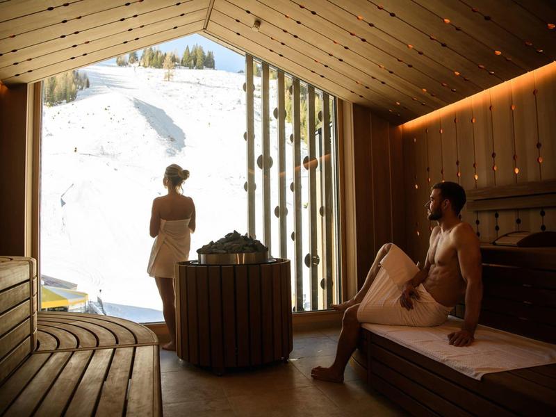 Ein Paar in einer Holzsauna mit Blick auf eine verschneite Landschaft durch ein großes Fenster.