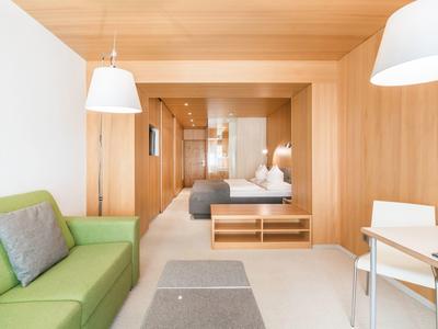 Helles Hotelzimmer mit grünem Sofa, Holzverkleidung und Doppelbett im Hintergrund.