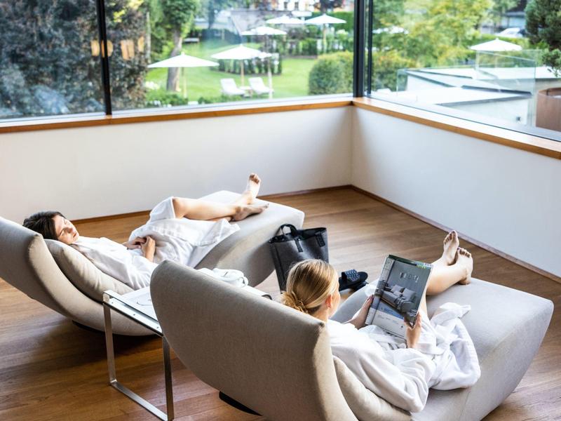 Zwei Personen entspannen in weißen Bademänteln auf modernen Sesseln mit Gartenblick.