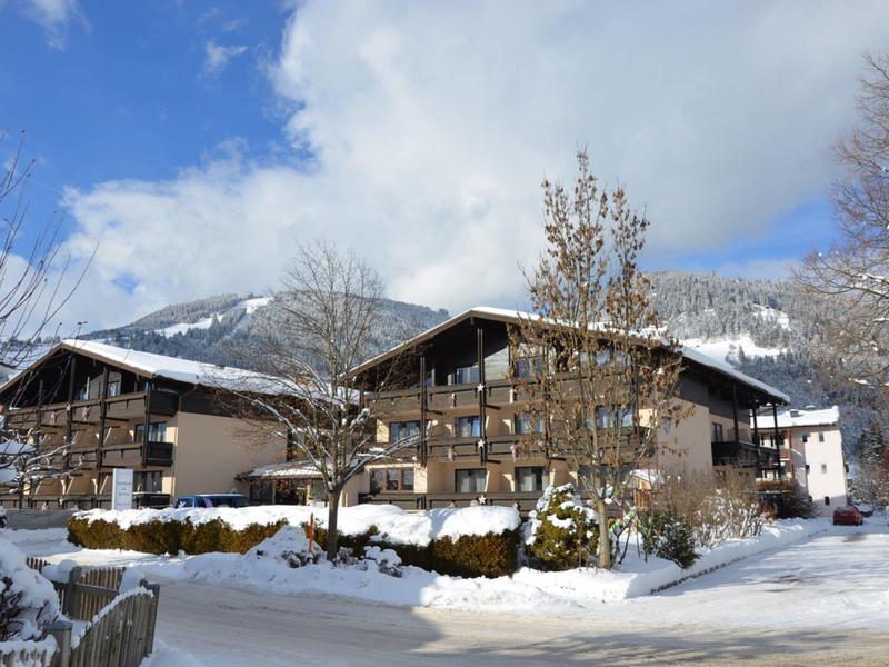 Zwei Chalet-Hütten in einem verschneiten Bergresort unter einem teils bewölkten Himmel.