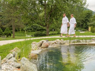 Zwei Personen in weißen Bademänteln gehen neben einem Teich in einem grünen Hotelgarten.