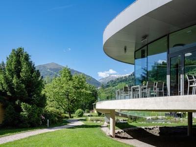 Modernes Hotel mit Glasfassade neben grüner Wiese und Bergen im Hintergrund.