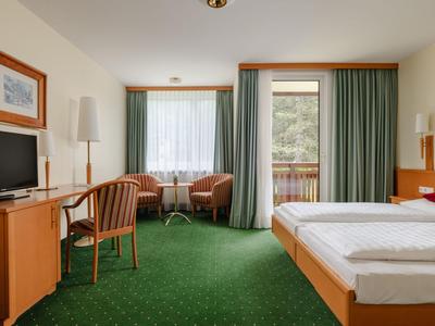 Helles Hotelzimmer mit grütem Teppich, Doppelbett, Schreibtisch, Fernseher und Balkon.