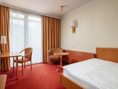 Helles Hotelzimmer mit Doppelbett, zwei Stühlen, Tisch und orangem Teppichboden.