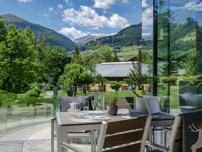 Terrasse mit weißen Tischen und Stühlen, Blick auf grüne Hügel und blauen Himmel.