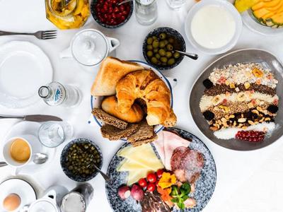 Verschiedene Frühstücksgerichte mit Croissants, Käse, Wurst, Obst und Müsli auf weißer Tischdecke.