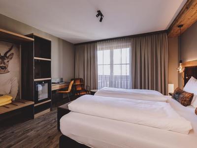 Hell eingerichtetes Hotelzimmer mit Doppelbett, Schreibtisch und großem Fenster mit Vorhängen.