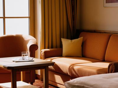Gemütliches Hotelzimmer mit oranger Couch, Sessel und Holztisch vor einem Fenster.