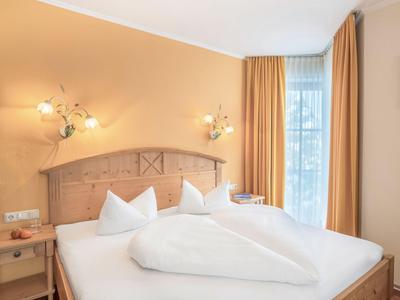 Camera d'albergo luminosa con letto matrimoniale, testiera in legno e tende color crema.