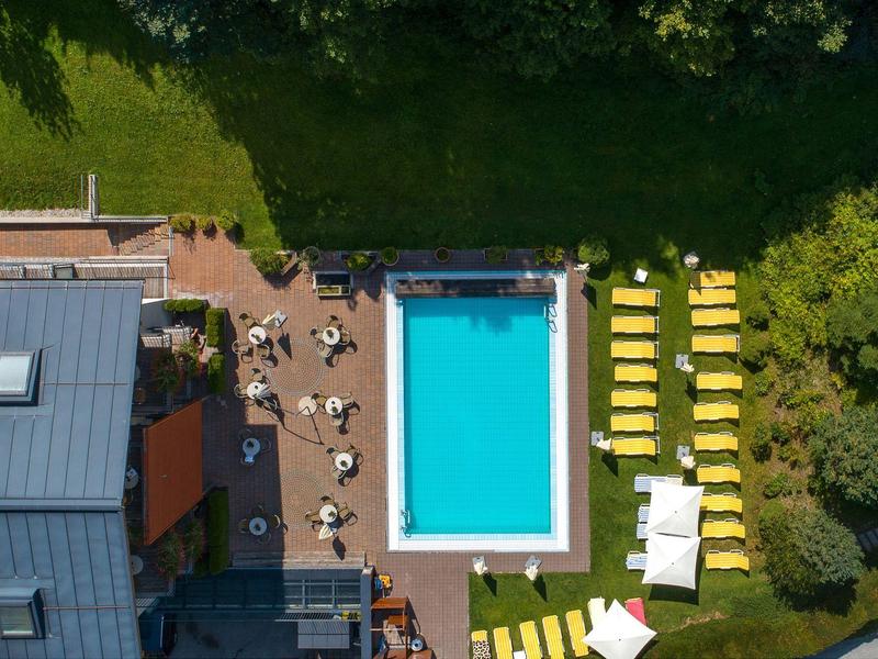 Rechteckiger Pool mit umliegenden Liegen und Tischen, daneben grüner Rasen und Gebäude.