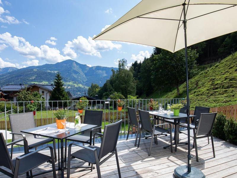 Terrasse mit Tischen, Stühlen und Sonnenschirm vor Bergen unter blauem Himmel.