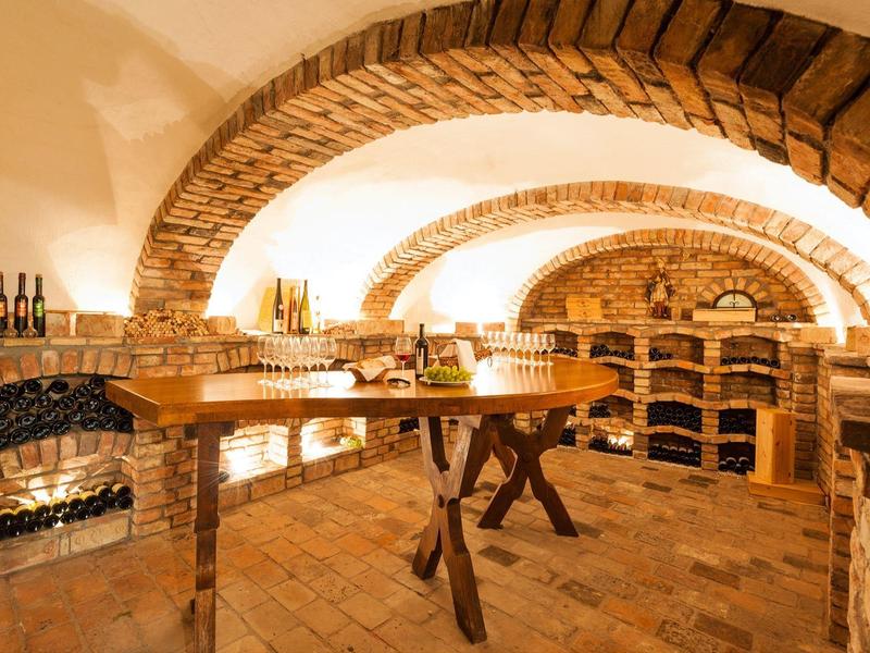 Gewölbter Kellerraum mit Ziegelbögen, Regalen für Weinflaschen und rustikalem Holztisch.