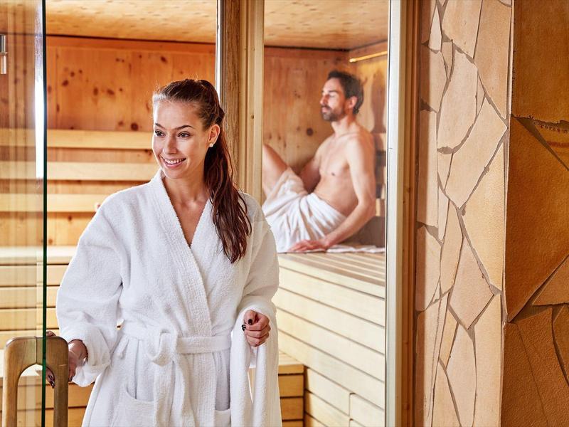 Frau im weißen Bademantel steht vor einer Holzsauna, zwei Männer sitzen im Saunaraum.