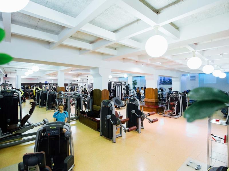 Großer, heller Fitnessraum mit vielen Trainingsgeräten, weißen Wänden und gelbem Boden.