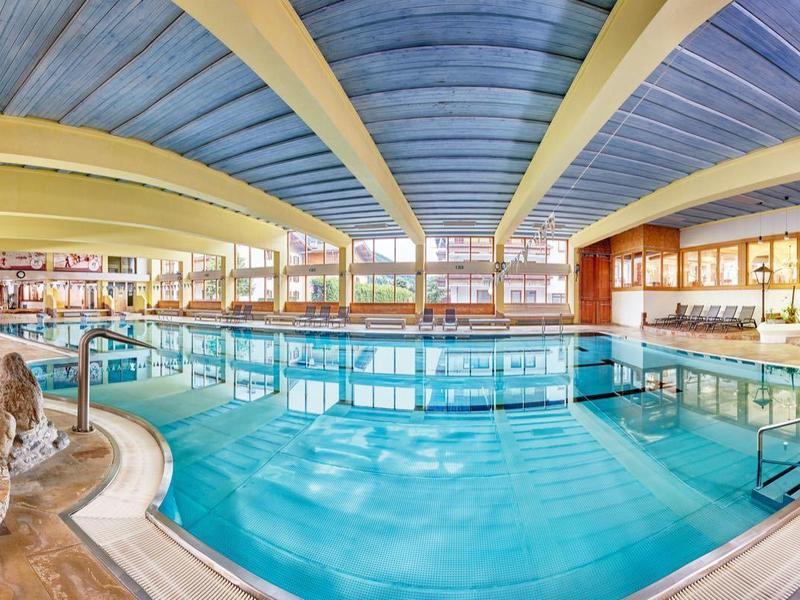 Innenbereich eines großen Hallenbads mit glattem blauem Wasser, Holzdecke und Panoramafenstern.