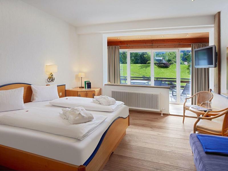 Helles Schlafzimmer mit Doppelbett, Holzmöbeln und großer Fensterfront mit Blick ins Grüne.