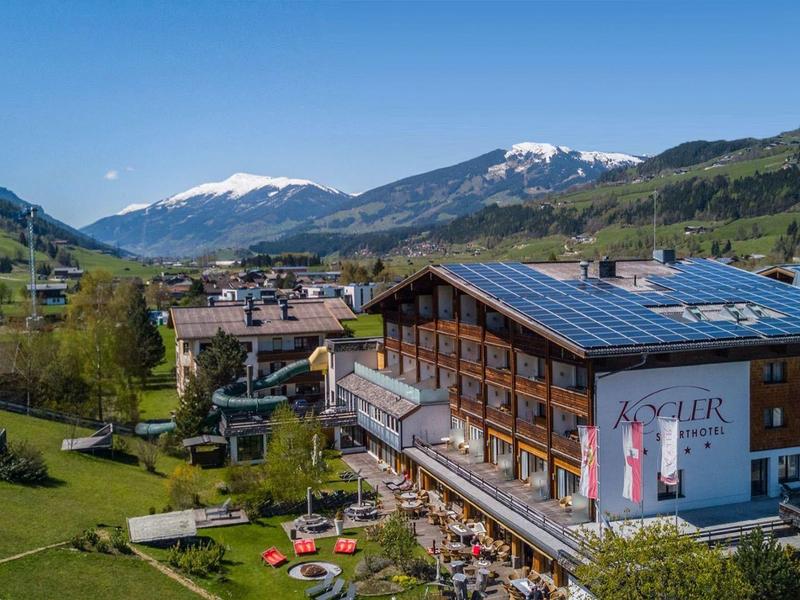 Bergdorf mit großen Holzhäusern, grünem Gras, Bergen und blauem Himmel im Hintergrund.