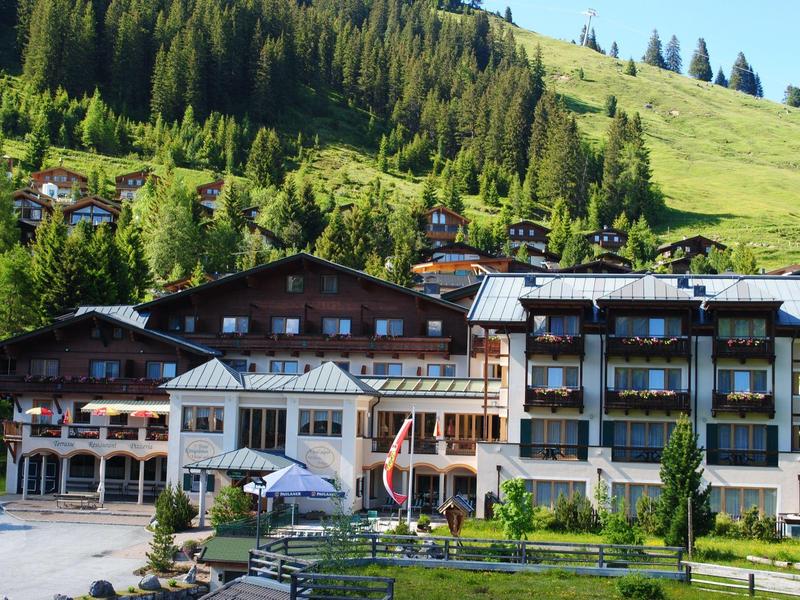Berghotel mit Holzbalkonen vor grünen Hängen und Bäumen an sonnigem Tag.