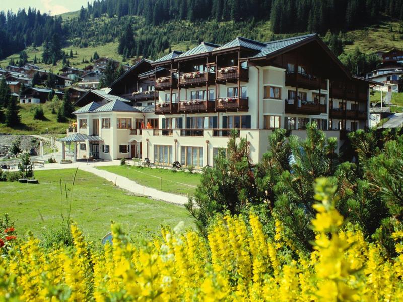 Alpenhotel vor grünen Wiesen, gelben Blumen und bewaldeten Bergen unter blauem Himmel.