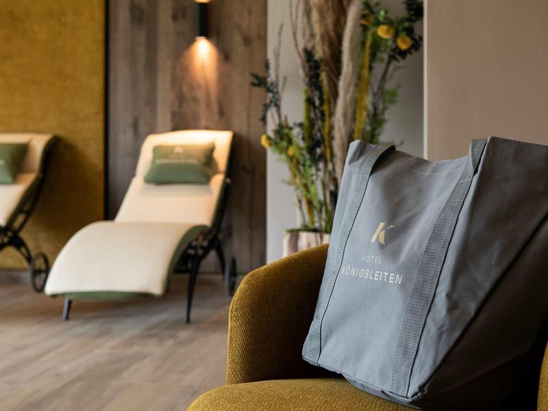 Moderner Wellnessraum mit zwei Liegen, Holzboden, großer Pflanze und grauer Tragetasche auf Stuhl.