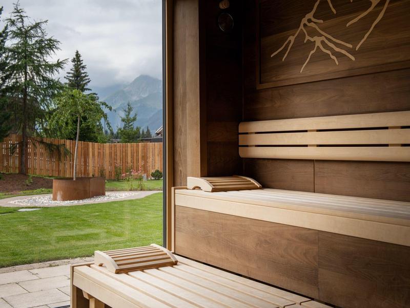 Holzsauna mit Liegen und Handtüchern, Blick auf grünen Garten mit Baum und Feuerstelle.