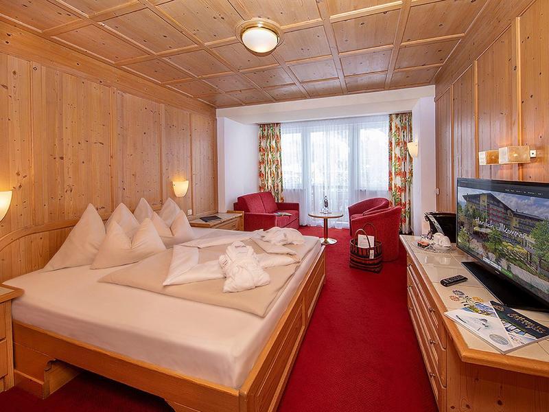 Gemütliches Hotelzimmer mit Holzverkleidung, rotem Teppich, großem Bett und Balkon im Hintergrund.