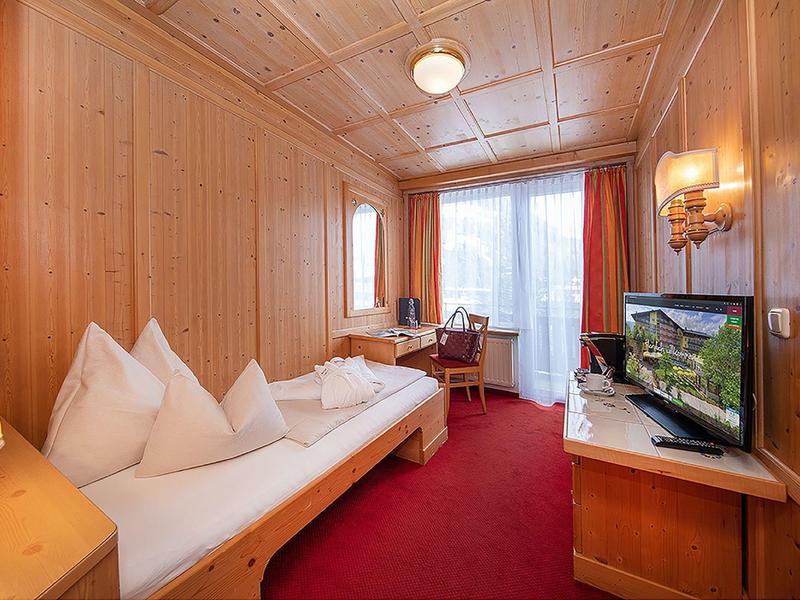 Kleines Hotelzimmer mit Holzwänden, rotem Teppich, Einzelbett, Balkon und Fernseher auf Kommode.