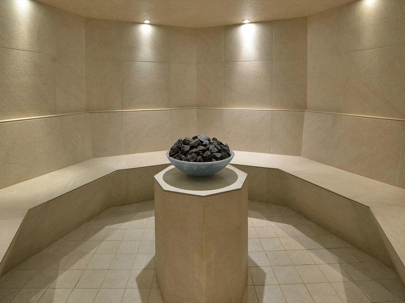 Runder, heller Sauna-Raum mit gebogenen Sitzen und einem Steintopf auf einem zentralen Podest.