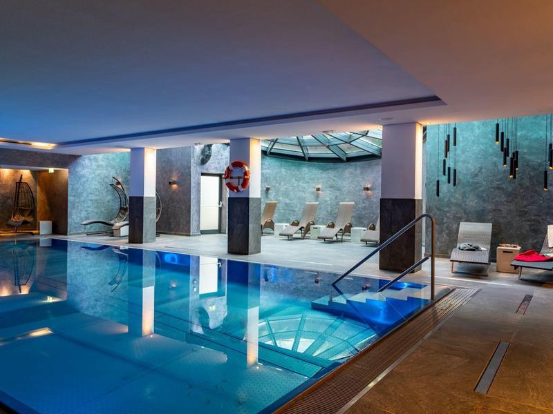 Innenpool mit bläulichem Wasser, Lounge-Bereichen, Liegestühlen und moderner Beleuchtung.