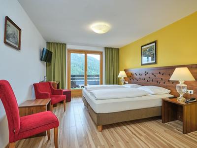 Světý hotelový pokoj s manželskou postelí, červenými židlemi, dřevěnou podlahou a výhledem na hory z okna.