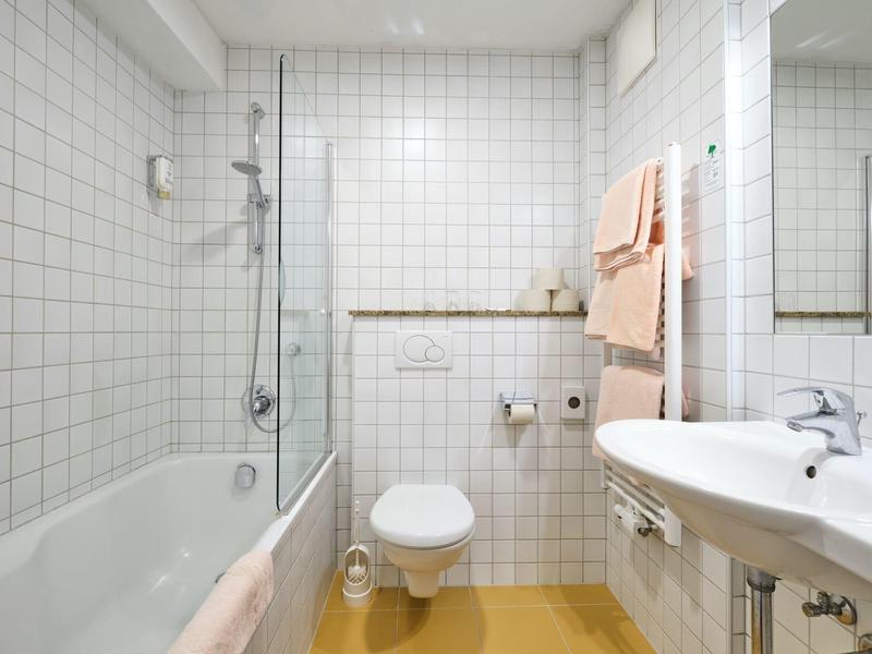 Kleines Badezimmer mit weißen Kacheln, Badewanne, Toilette und Waschbecken, Handtücher an der Wand.