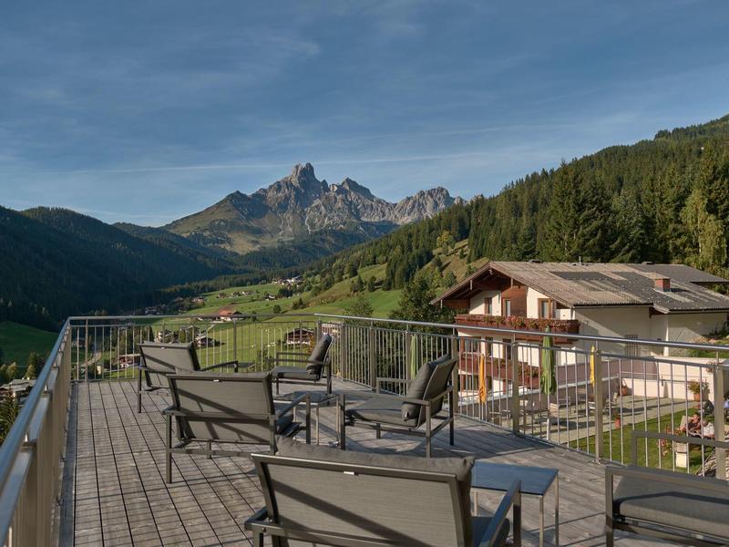 Bergterrasse mit Sitzmöbeln, Blick auf bewaldete Hügel und markante Bergspitzen im Hintergrund.
