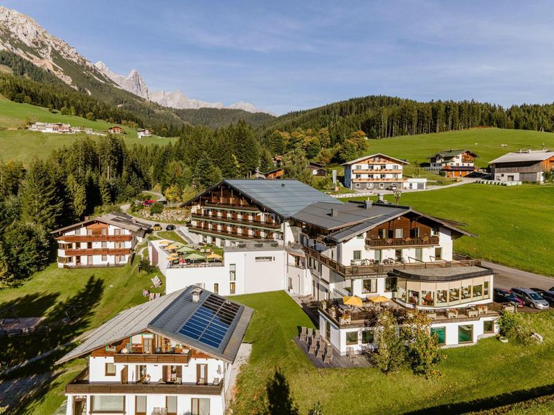 Bergdorf mit Holzhäusern und Wiesen, umgeben von Wäldern und Bergen unter klarem Himmel.