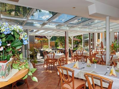 Bright restaurant with glass roof, wooden tables and chairs, decorated with flowers.