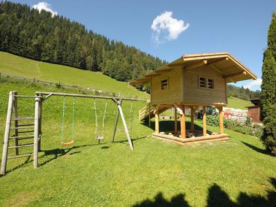 Spielplatz mit Schaukel und Baumhaus auf einem grünen Rasen mit Wald im Hintergrund.