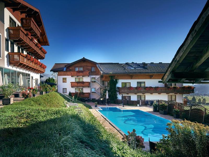 Blick auf ein Hotel mit Außenpool, weißen Wänden, Holzbalkonen und grünem Rasen unter blauem Himmel.