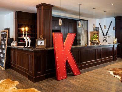 Réception d'hôtel avec comptoir en bois foncé, grande lettre "K" et éléments décoratifs.