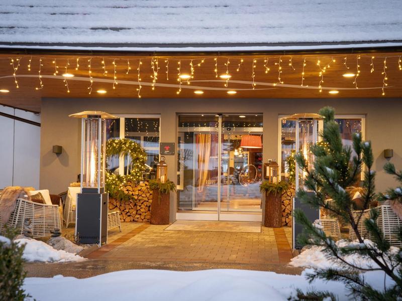 Eingang eines Hauses mit Glastür, beleuchtet von warmen Lichterketten und mit Schnee im Vordergrund.